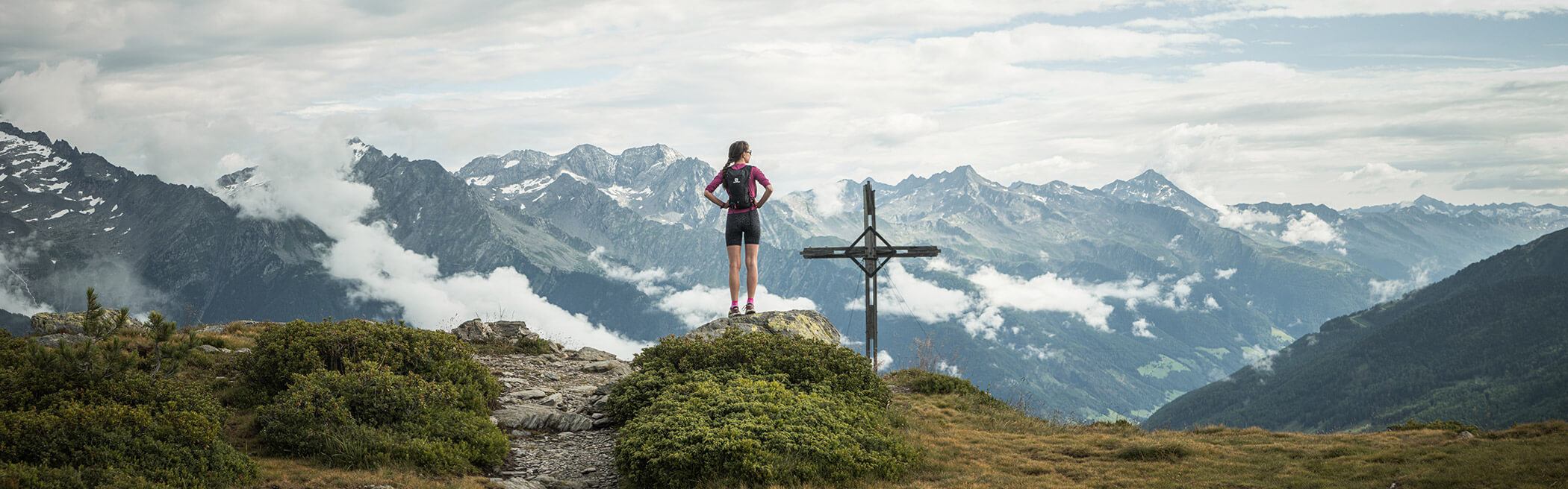 Junge Frau steht beim Gipfelkreuz, hinter ihr ein monströses Bergpanorama - OLM Nature Escape