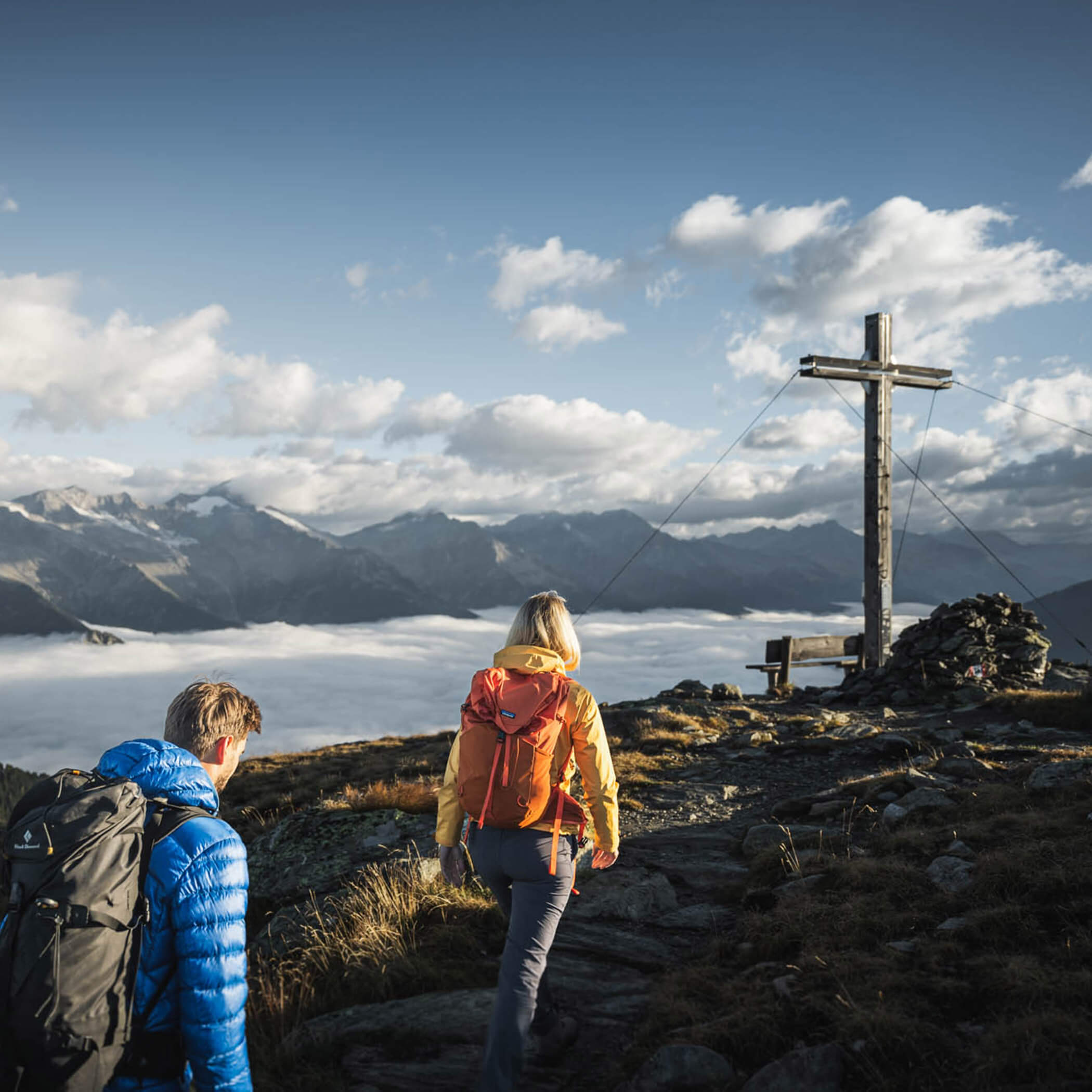 Wanderer erreichen ein Gipfelkreuz mit Bergpanorama im Naturpark Rieserferner-Ahrn - OLM Nature Escape