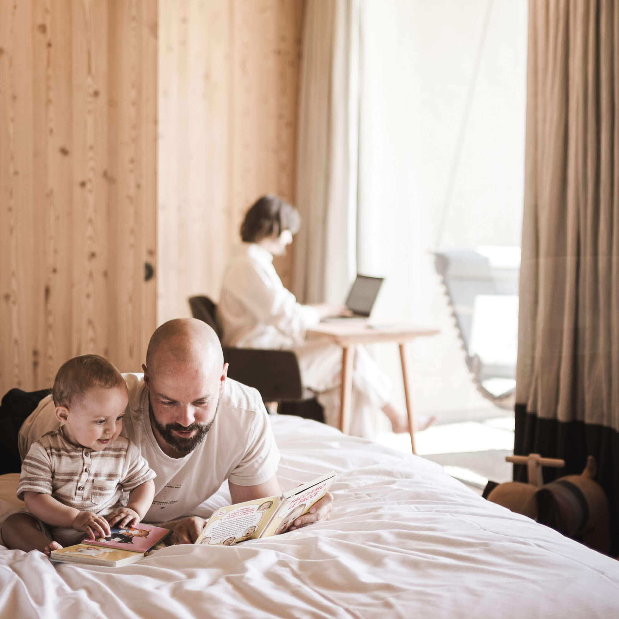 Vater und Sohn lesen ein Buch, Mutter im Hintergrund arbeitet am Laptop